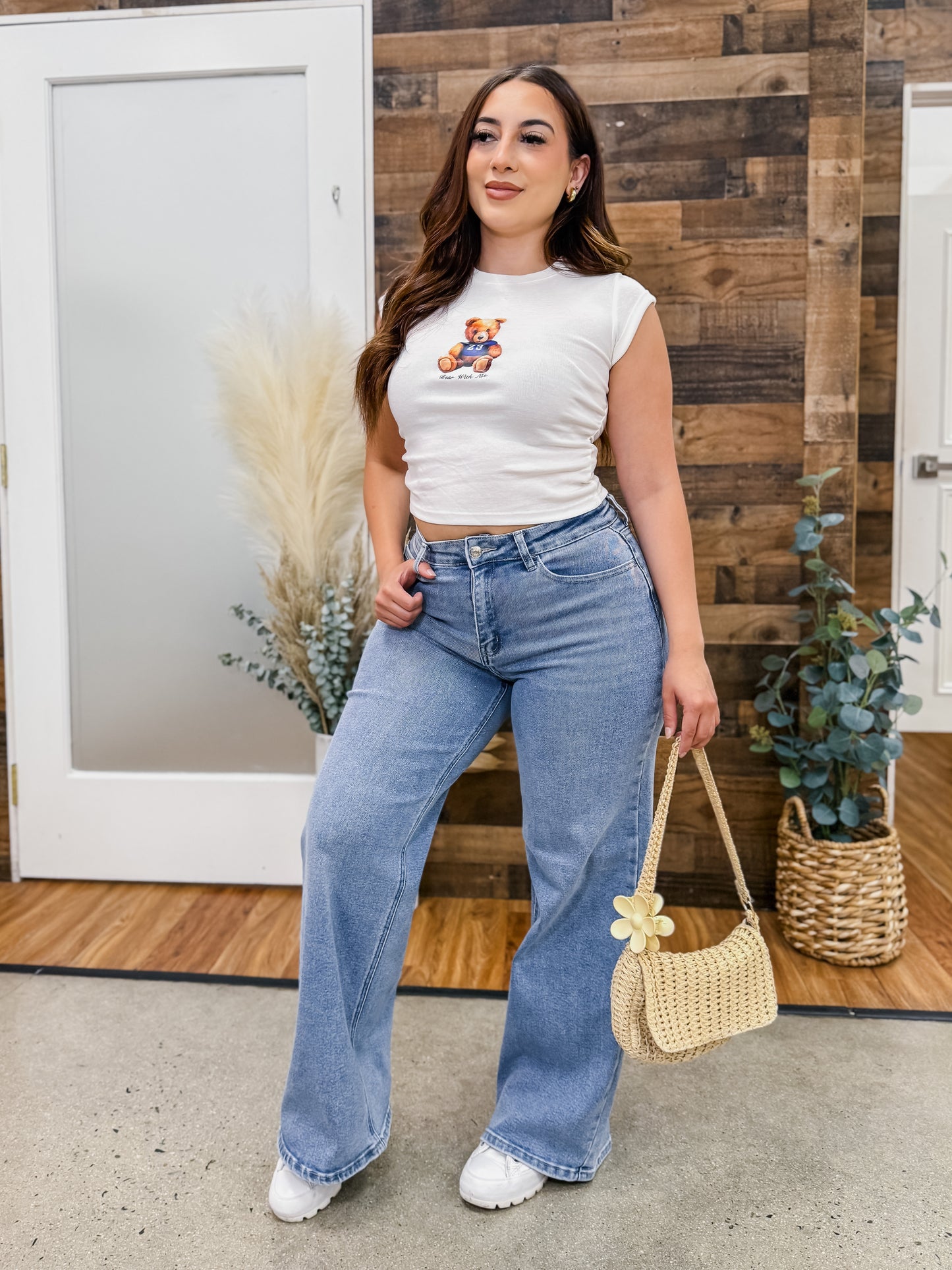 Woman wearing a white crop top with a teddy bear design and blue jeans, standing in front of a wooden wall.