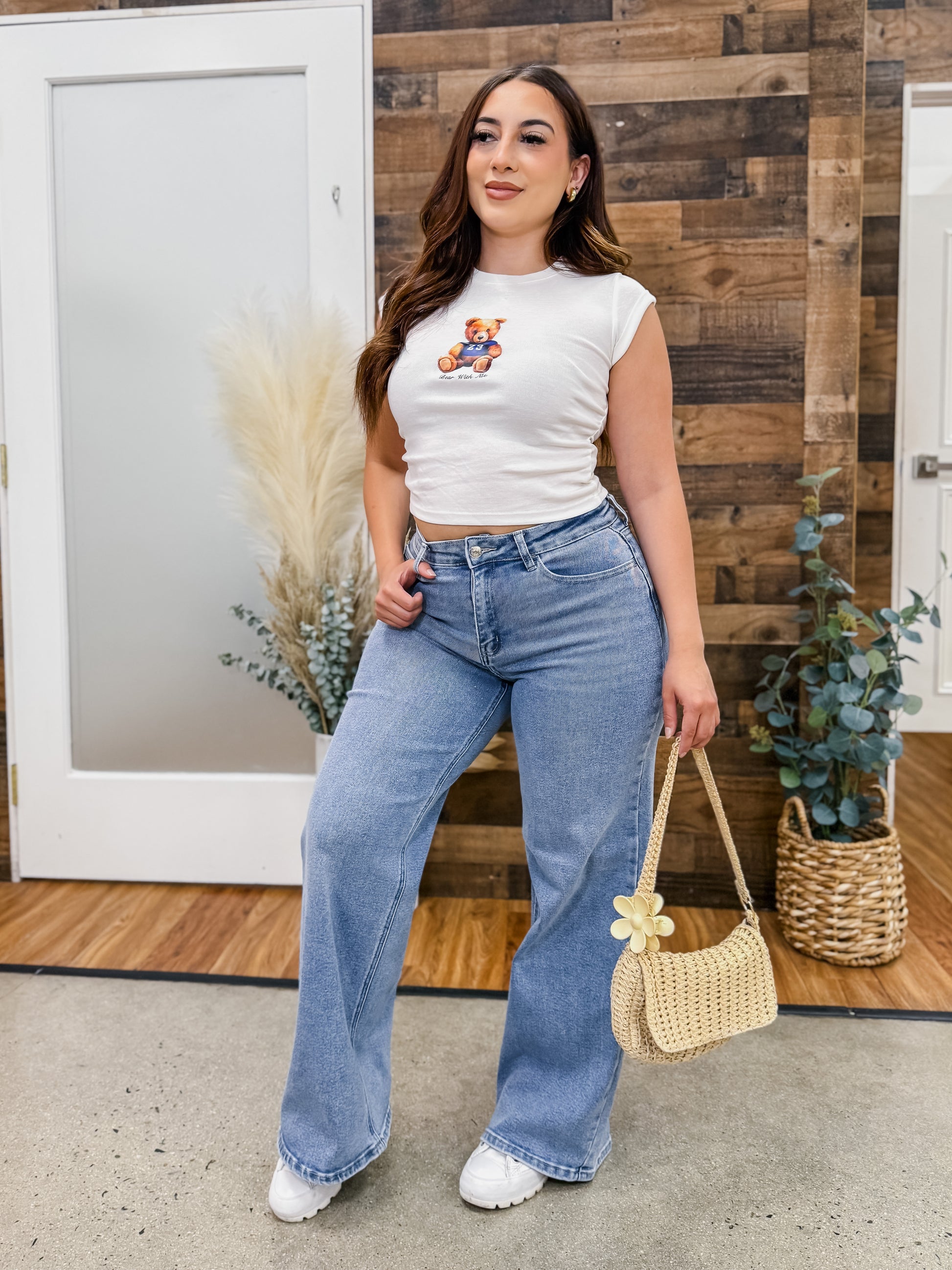 Woman wearing a white crop top with a teddy bear design and blue jeans, standing in front of a wooden wall.