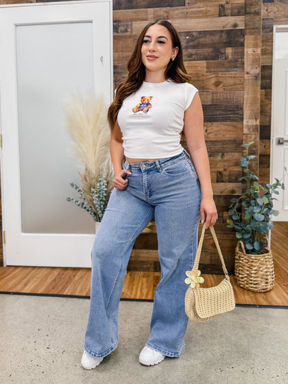 Woman wearing a white crop top with a teddy bear design and blue jeans, standing in front of a wooden wall.