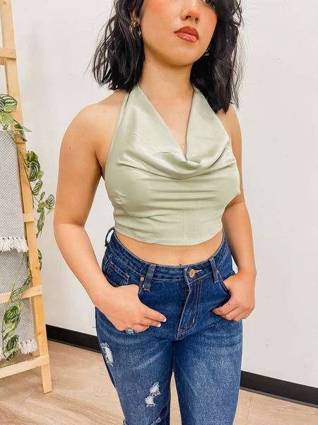 Close up picture of short black haired model wearing Lucia Top in sage green color. She has her hands in the pockets of dark wash jeans.