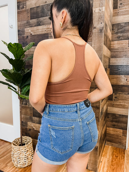 Model is turned arround to show the raceerback style of the brown bodysuit. She is wearing medium wash jean shorts.