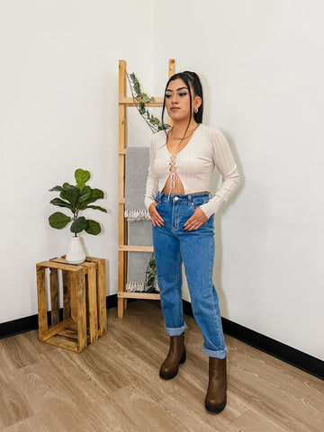 Woman with ponytail wearing a long sleeve top and blue jeans with plant in background