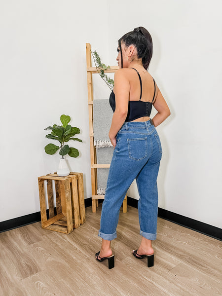 Full body picture of the model turning her back to the camera. The model is wearing black shirt with medium wash blue jeans and low black heels. SHe is standing in front ofwooden ladder and wooden crate with plant on top.