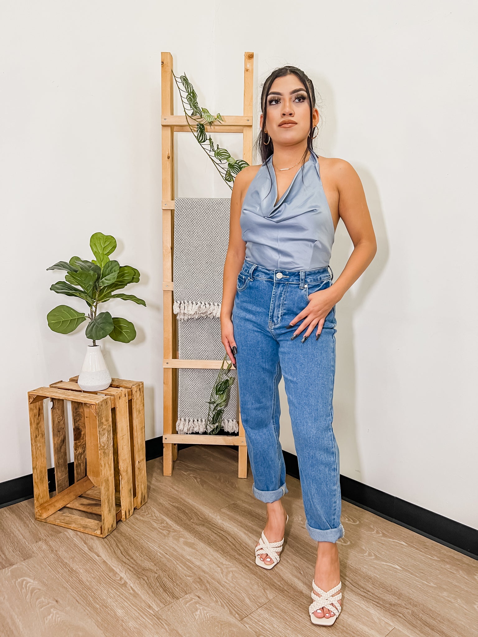 Model is standing in front of white wall with wooden ladder and crate decorated with plants. She has her hair up in a pony tail wearing folded hem jeans and a light blue silk sleeveless top. She is also wearing braided white heels. 