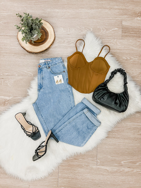 Flatlay image of corset brown top with black small purse to the right. The clothing is placed on top of a white rug against light brown wooden floor. To the left of the top light wash jeans are folded with earrings on top with low black heels . Above the outfit is a small plant.